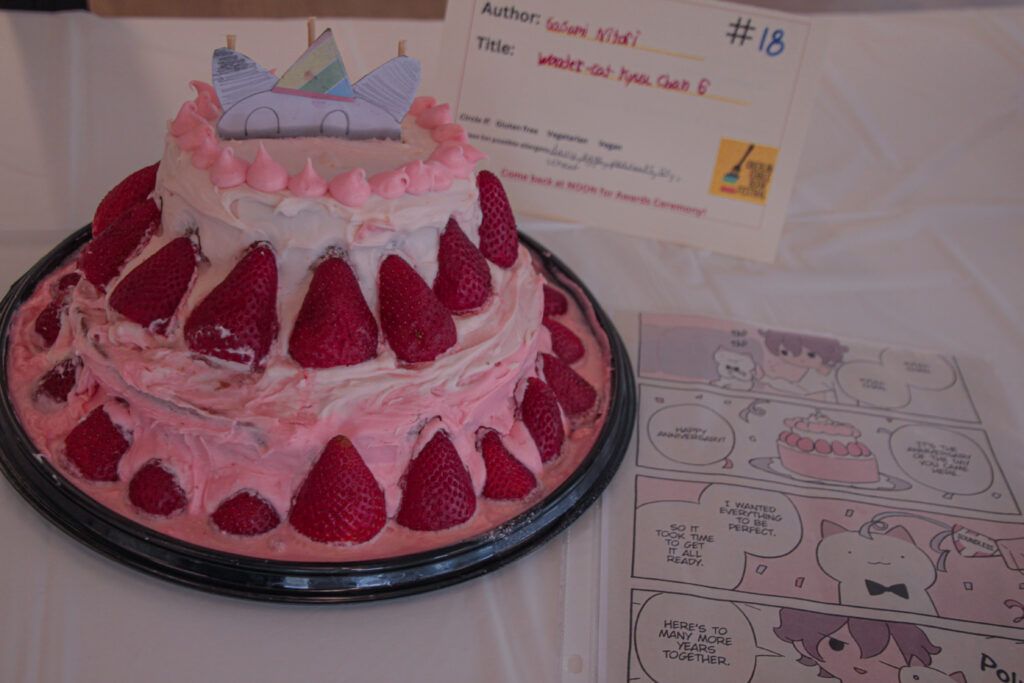 Pink frosted cake with strawberries and a paper illustrated cat head poking out of the top, accompanied by a page of the piece it's referencing.
