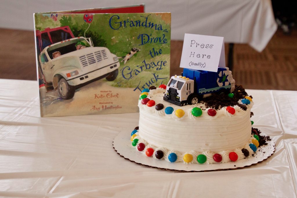 Photo of submission with a cake featuring a toy garbage truck on top with a card reading "Press Here (Gently)" in front of the book "Grandma Drove the Garbage Truck" by Katie Clark
