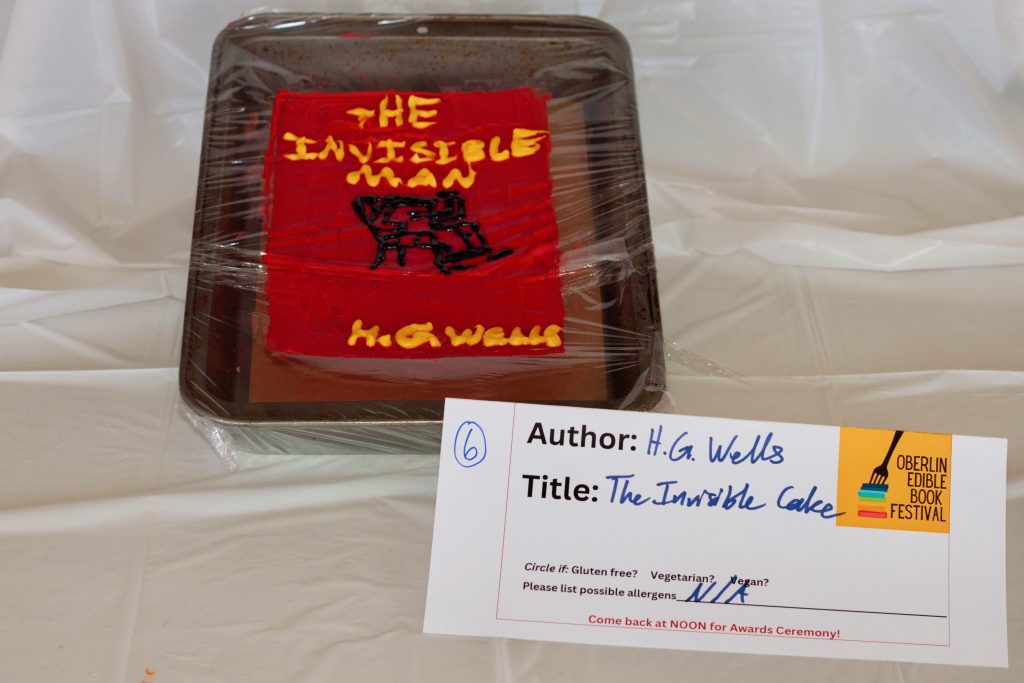 Photo of submission titled, "The Invisible Cake" featuring a red cake with the title of the book on it and wrapped in plastic wrap